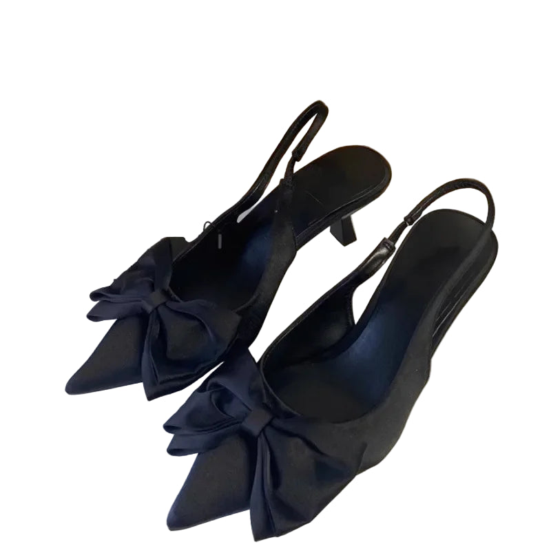 Black high-heeled shoes with bow details on a white surface, next to a cardboard box.